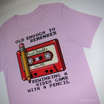 A pink t-shirt. It has a red tape cassette with cream / white lable. Text reads "old enough to remember rewinding a video game with a pencil"